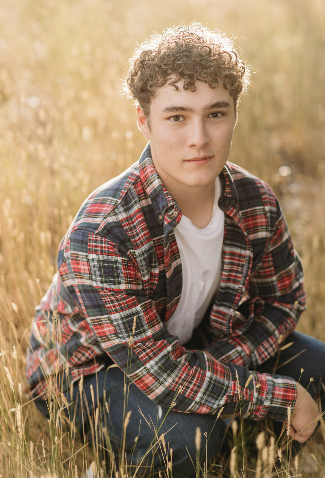 a boy sits in a field looking at the camera wearing a plaid shirt